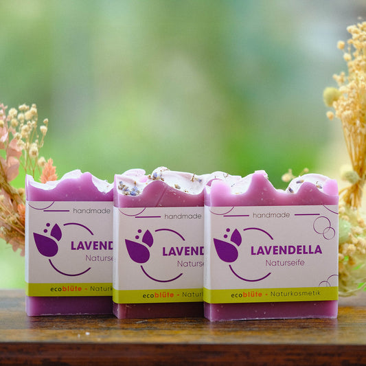 Naturseife Lavendella mit Banderole, handgemacht in unserer Seifenmanufaktur, eine ideale Körper- und Gesichtsseife strahlend in Laveldelfarbe, Gesteck im Hintergrund, 3er Set