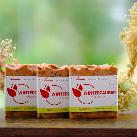 Naturseife Winterzauber mit Banderole, handgemacht in unserer Seifenmanufaktur, eine Peelingseife die nach Weihnachten duftet, Gesteck im Hintergrund, 3er-Set