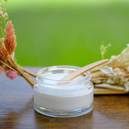 Deo-Creme Tiegel offen mit Spatel und Gesteck im Hintergrund